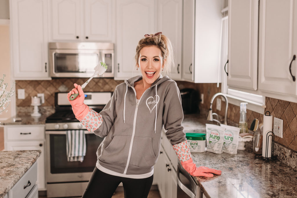 Brianna K bitsofbri in her kitchen sharing 5 mom hacks for around your home in her blog post holding a dish scrubber and using Cascade Pure Essentials 