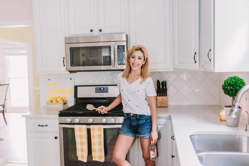 Brianna K wearing white tee shirt with black stars and blue jean cut off shorts with an orange scarf tied around her hair sharing how to clean your stove with natural cleaning products in her new all white kitchen decorated with pops of yellow decor for summer 2019.