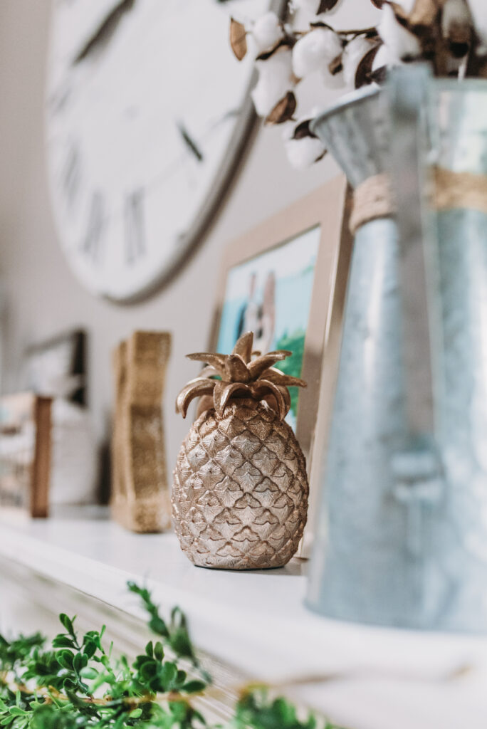  mantle decor ideas for summer- blue candle holders, gold pineapple, white pineapple, cotton filled vase, large white clock hanging able mantle with family pictures and greenery garland strung across mantle. Bitsofbri Brianna K summer decor home tour 2019 blog post summer decor inspiration 
